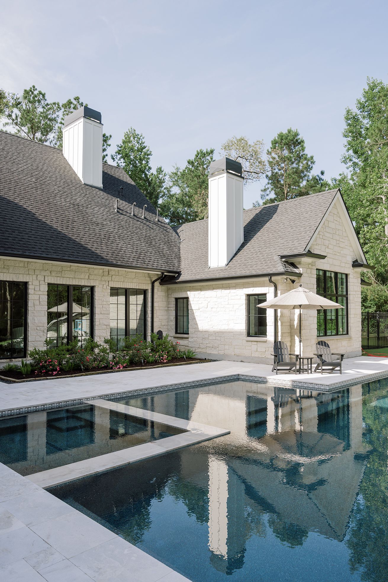 Luxury home exterior with white chimney, painted brick, black windows, and rectangular pool with patio seating.