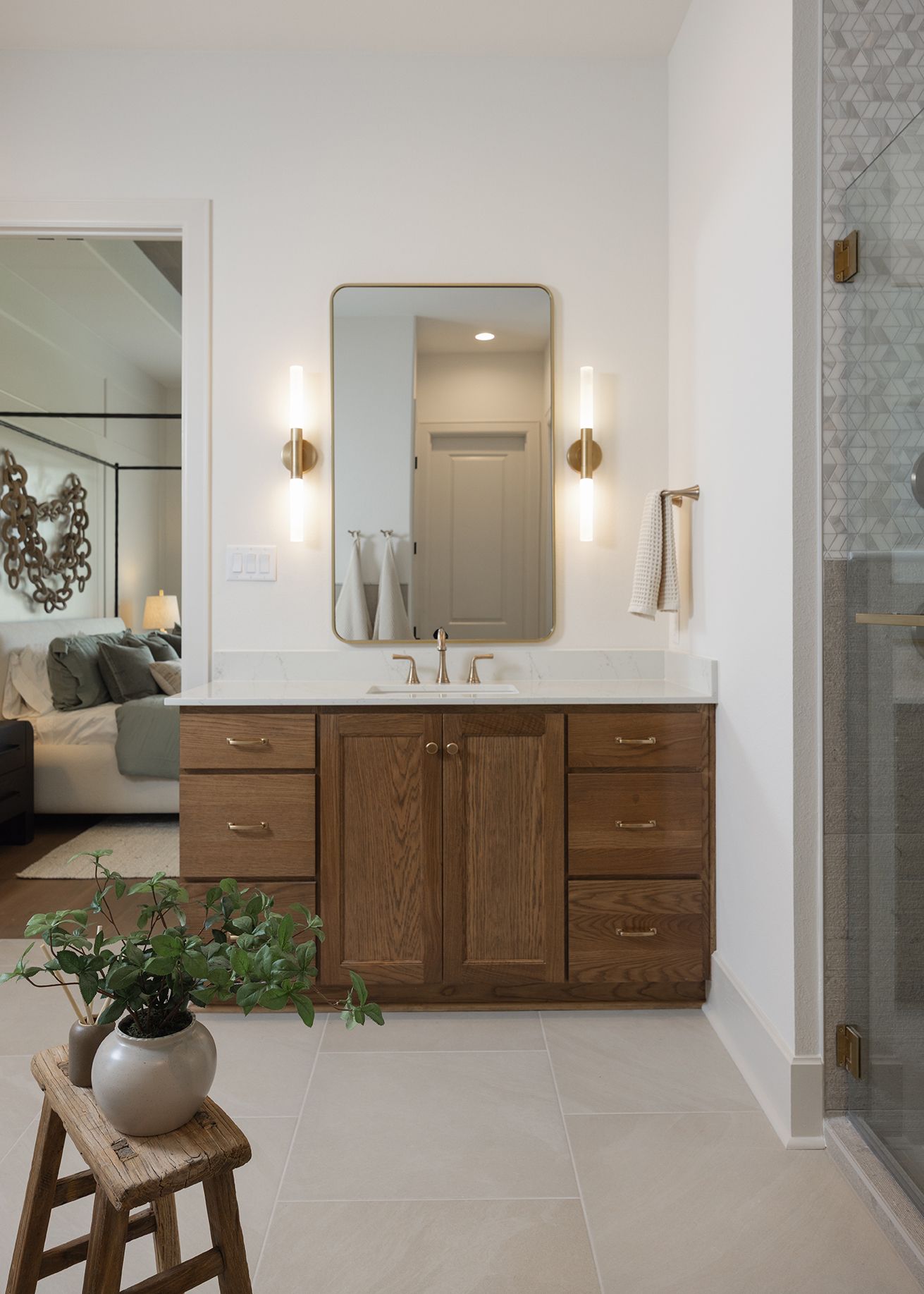 Elegant bathroom vanity with warm wood cabinets, brass fixtures, rounded mirror, and gold sconce lighting beside a glass shower.