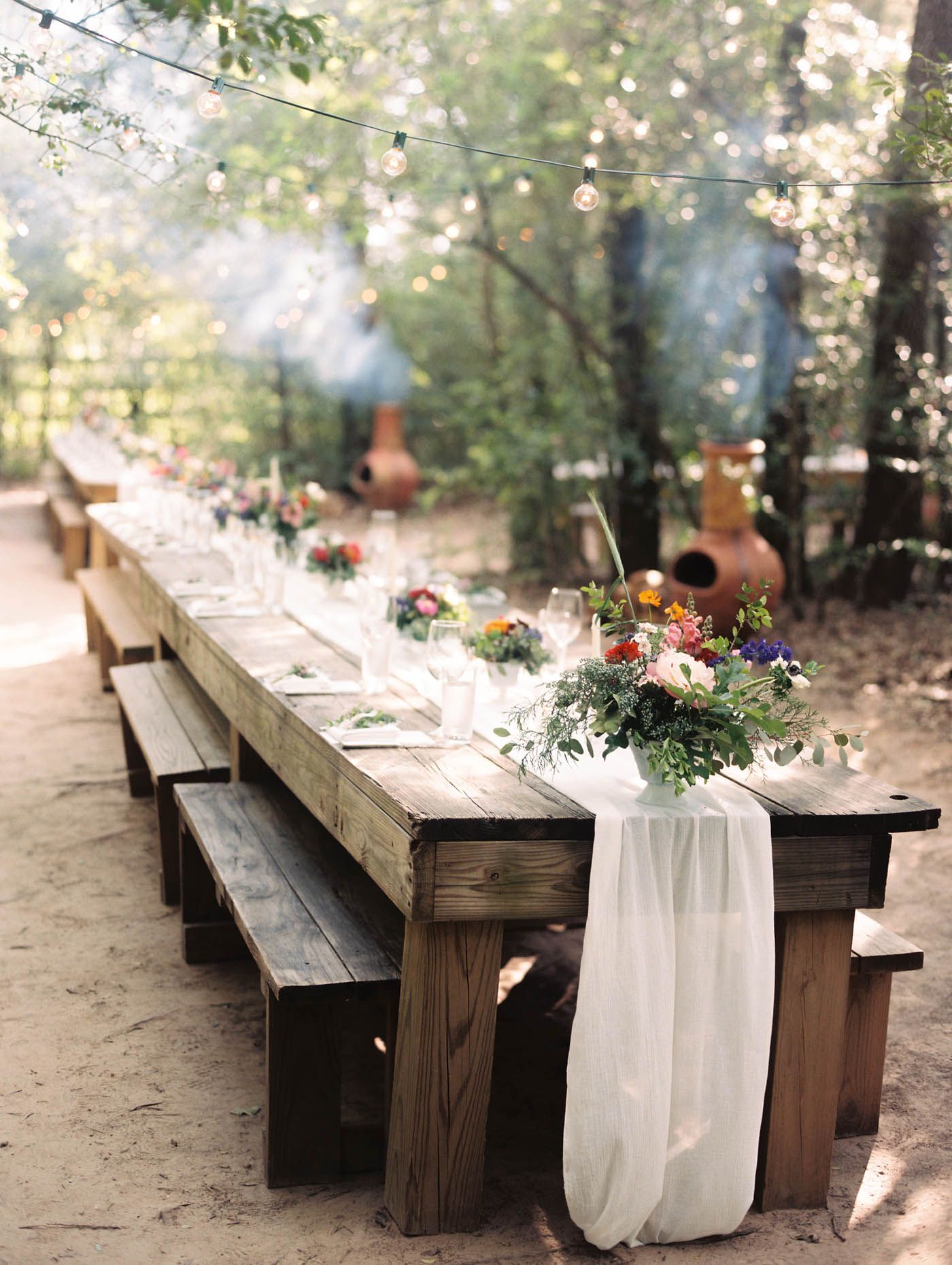 Rustic outdoor wedding reception with long wooden farm table, white chiffon runner, colorful floral centerpieces, and string lights.