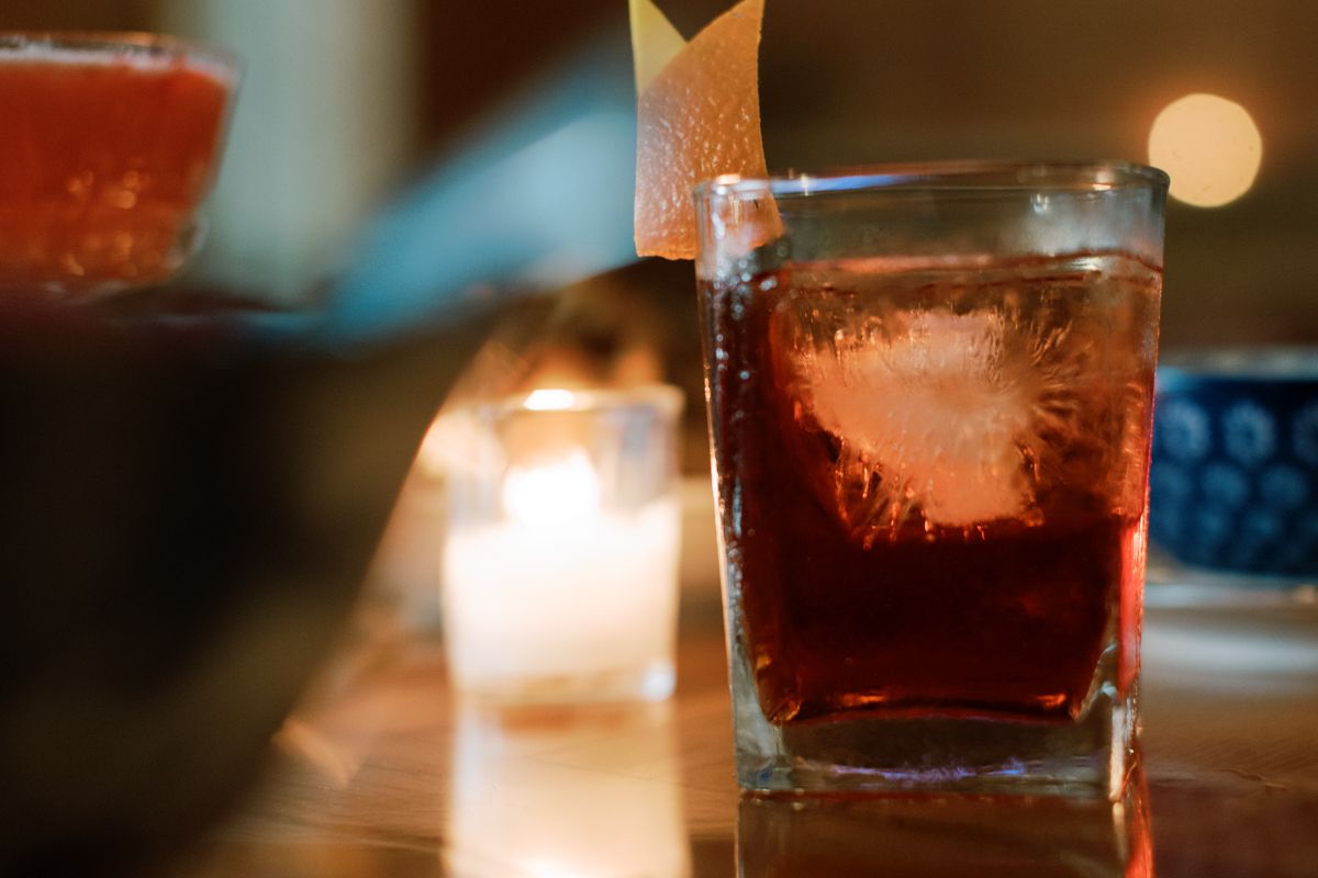 A dark whiskey cocktail with ice and citrus garnish in a rocks glass, with candles glowing softly in the background.