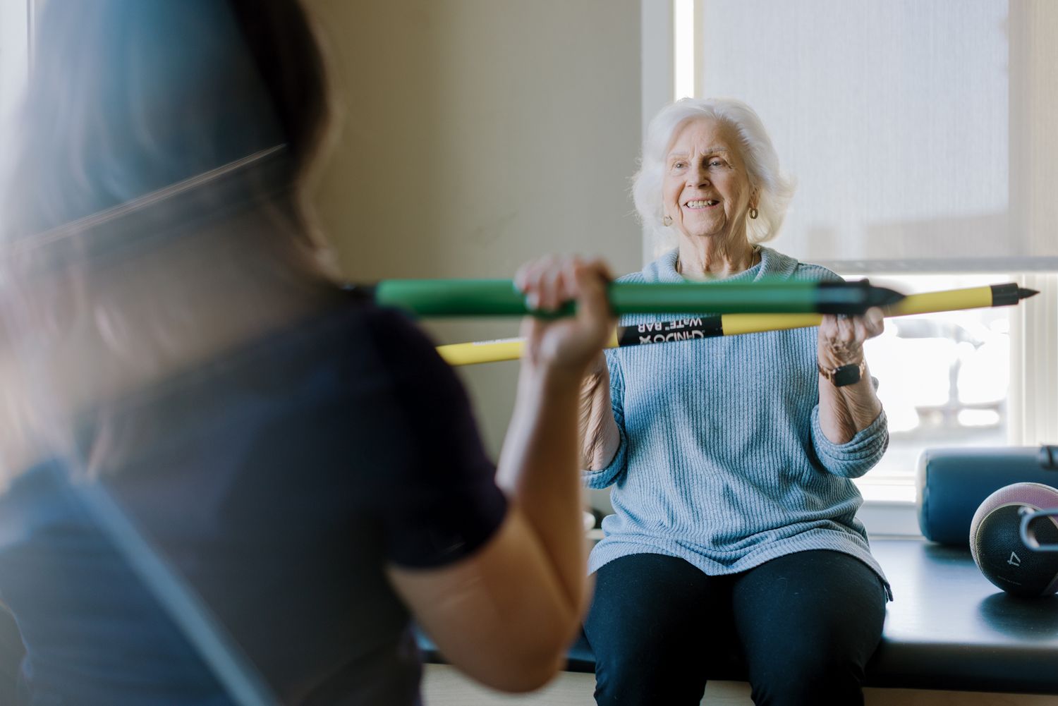 Senior woman smiling while exercising with a bar during a fitness session with a trainer.