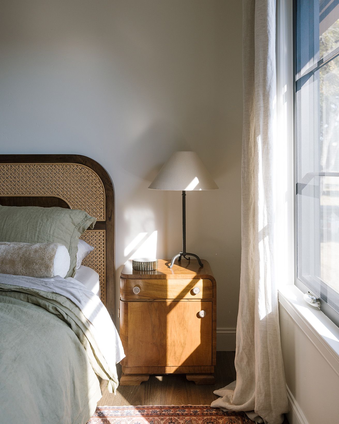 Serene bedroom corner with rattan headboard, vintage wood nightstand, white lamp, and linen curtains.