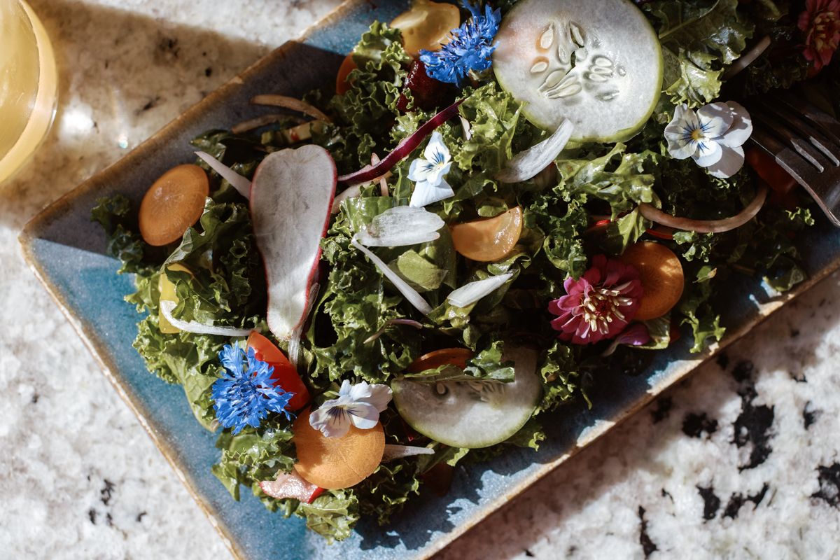 Fresh kale salad with edible flowers, cucumber, cherry tomatoes, and radishes on a blue ceramic plate.