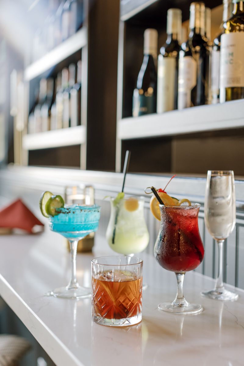 Five colorful cocktails on a white bar counter with bottles on shelves behind.