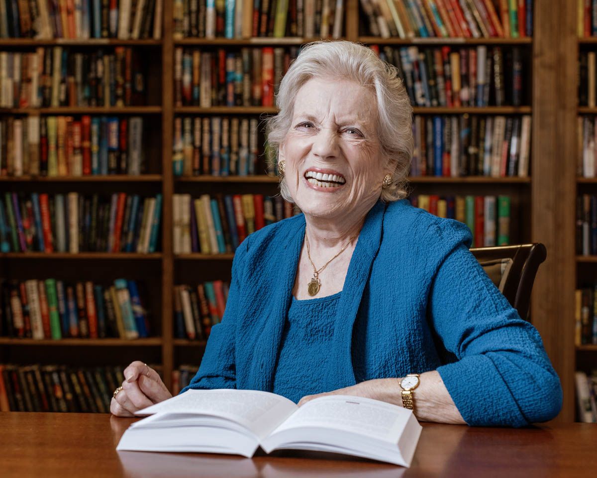 Happy elderly woman laughing while reading an open book in a library filled with colorful bookshelves.