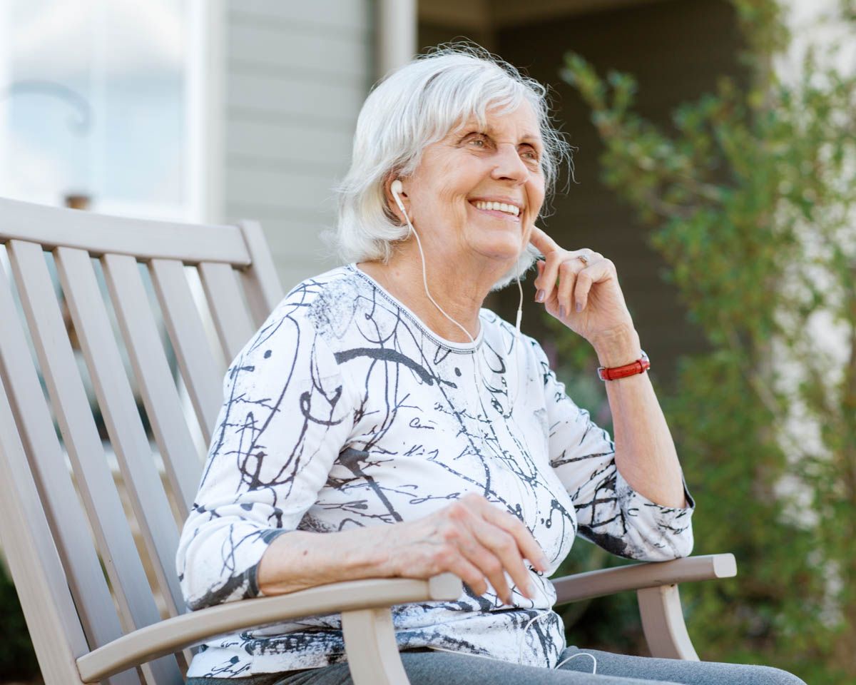 Smiling senior woman with white hair sitting outdoors on a bench, enjoying music with earbuds on a sunny day.