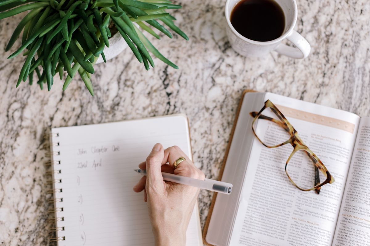 A hand writing in a spiral notebook beside an open book with glasses, a coffee cup, and a green plant on a marble surface.