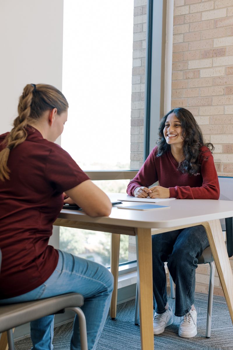 Two college students smile and chat at a table near a bright window on campus.
