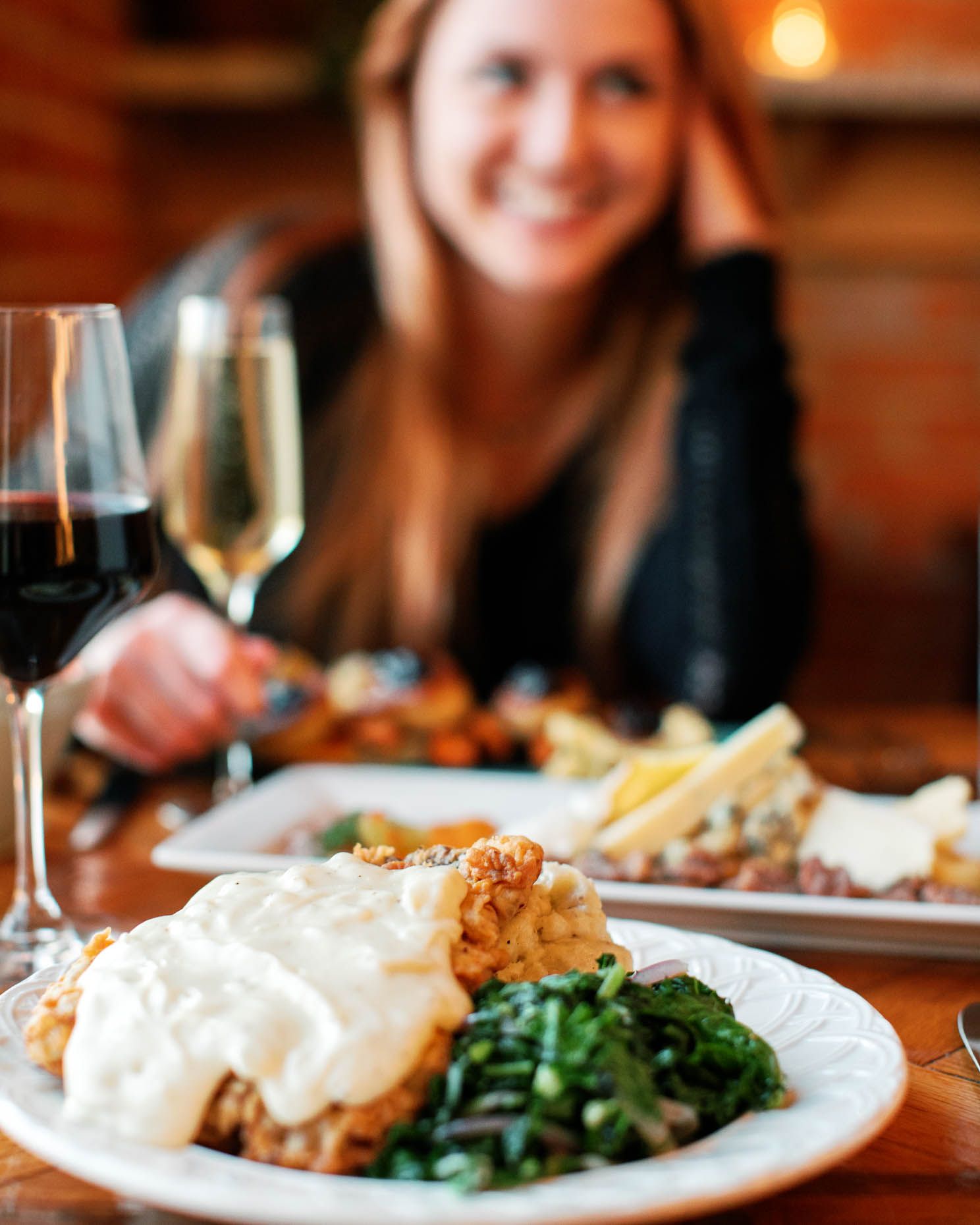 Woman smiling at a restaurant table with a white plate of chicken and greens and two glasses of wine.