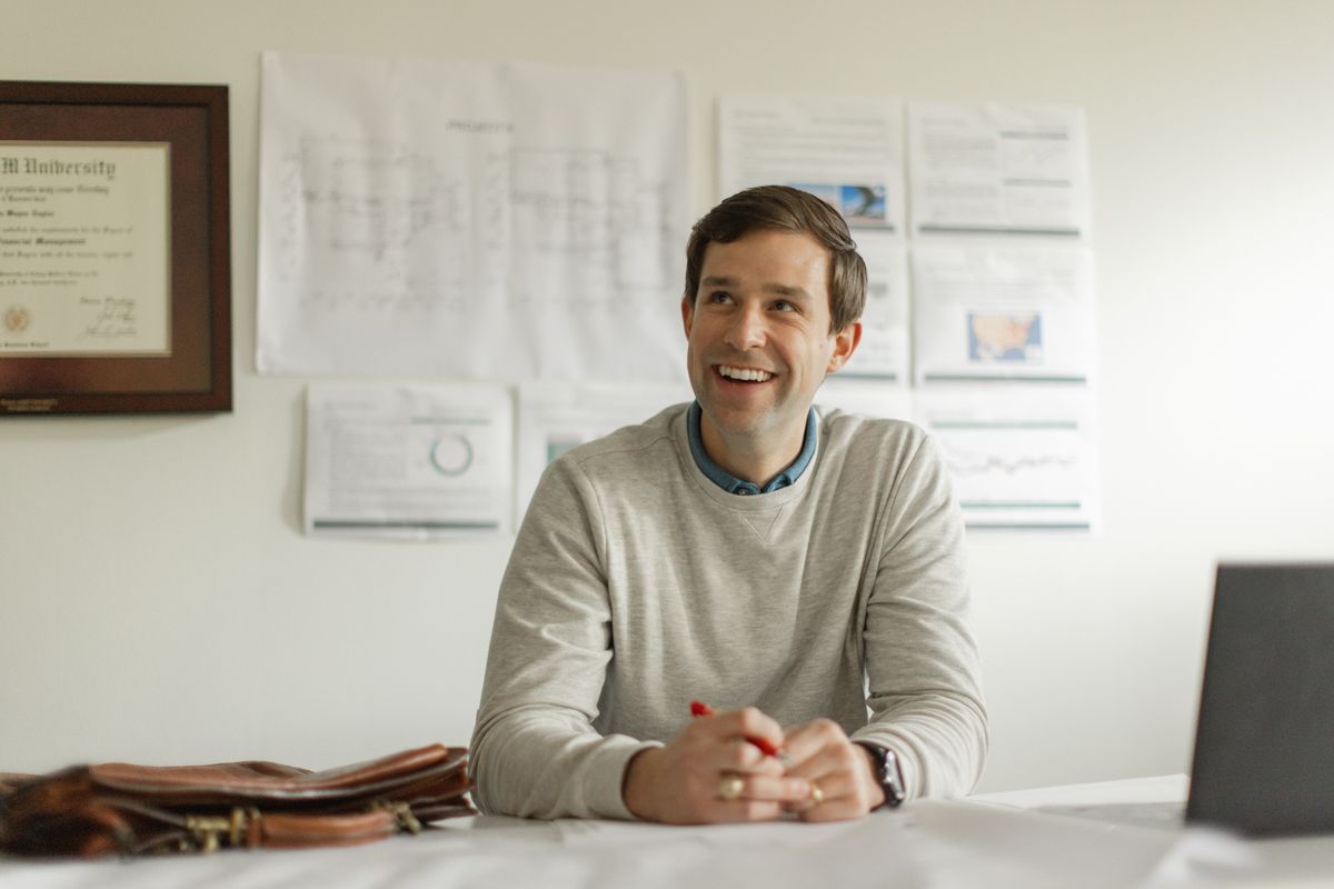Smiling professional man sitting at desk with blueprints and charts on wall behind him in modern office.