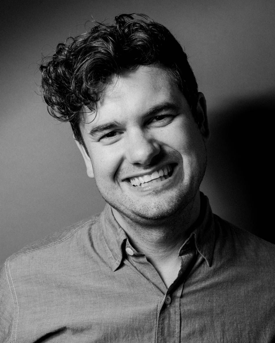 Black and white portrait of a smiling young man with curly hair wearing a button-up shirt.