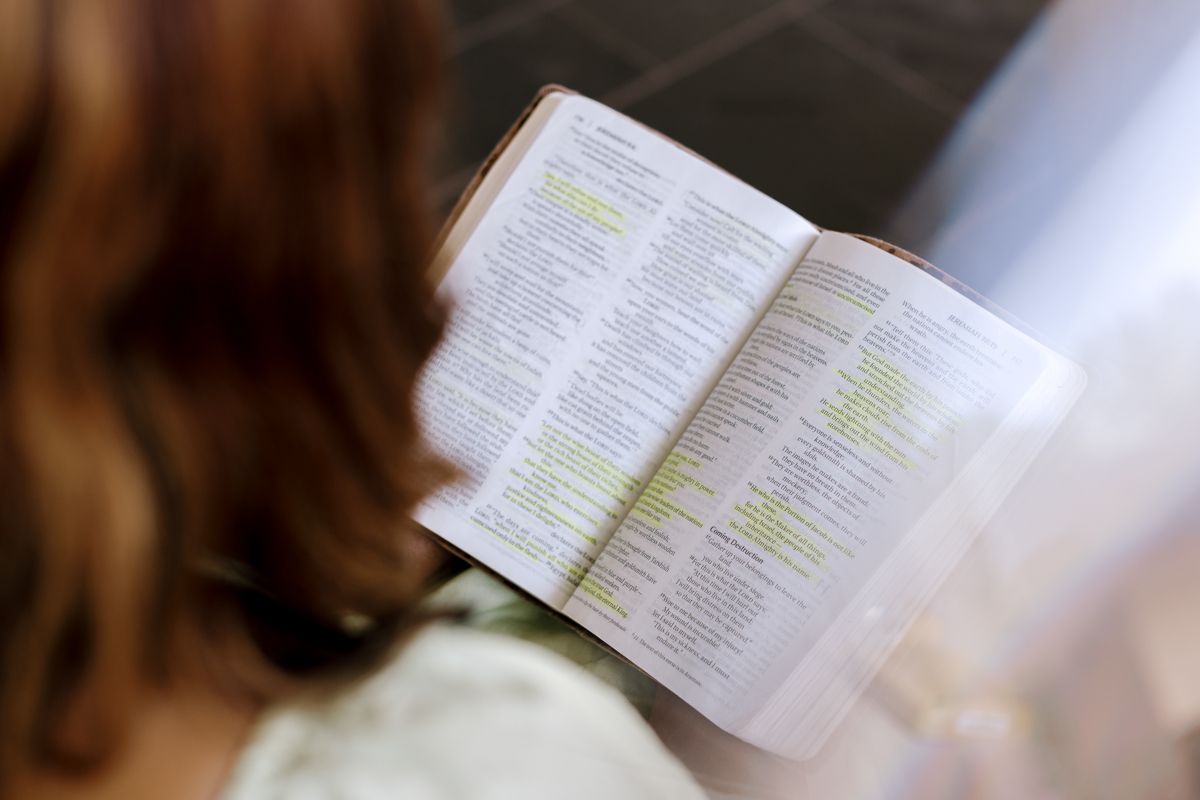 Woman reading a highlighted Bible or book, viewed from behind in a bright indoor setting.