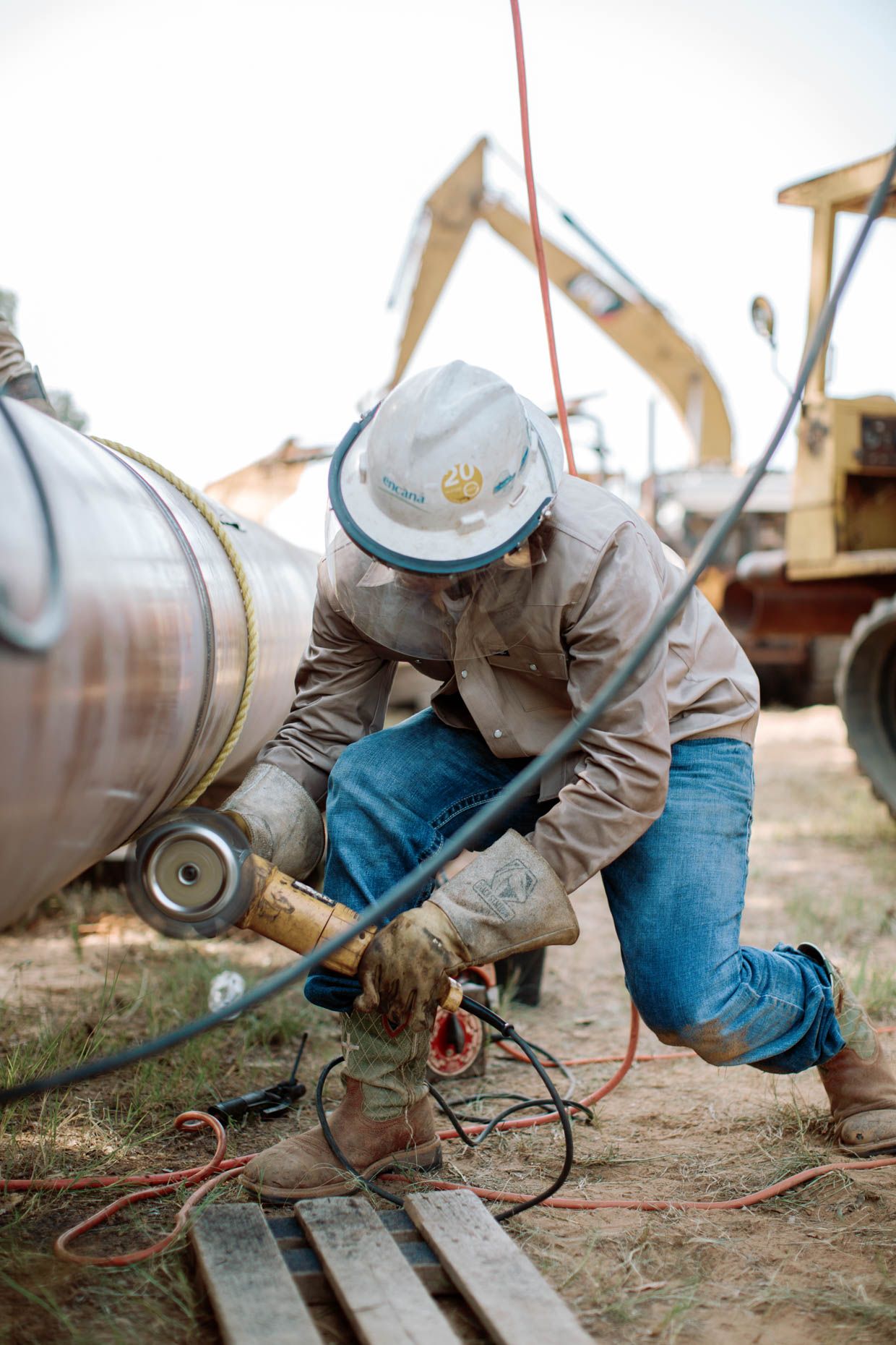 Construction worker in hard hat kneeling to connect fittings on large industrial pipe at job site.