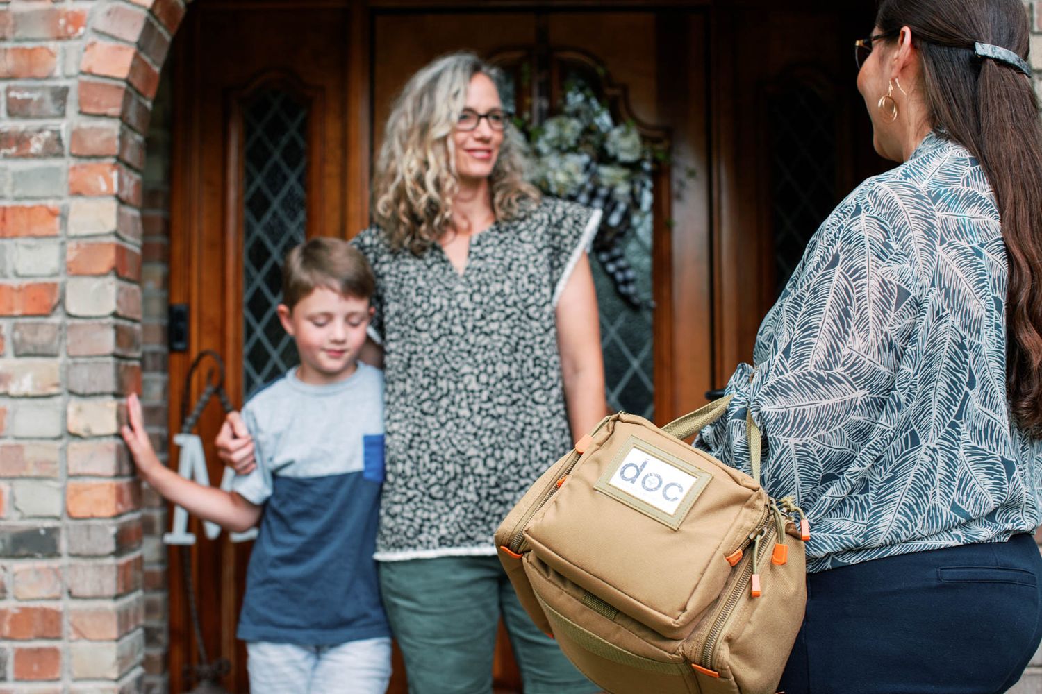 A woman and child greet a visiting healthcare provider with a medical bag at their front door.
