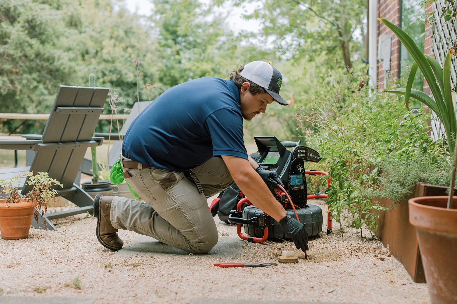 Technician in navy shirt kneeling outdoors using diagnostic equipment on a patio near plants.