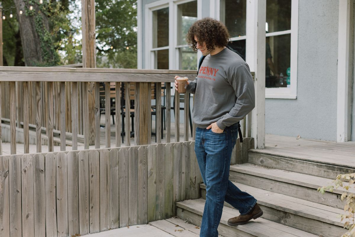 Curly-haired man in Penny sweatshirt walks down wooden porch steps holding a coffee cup.