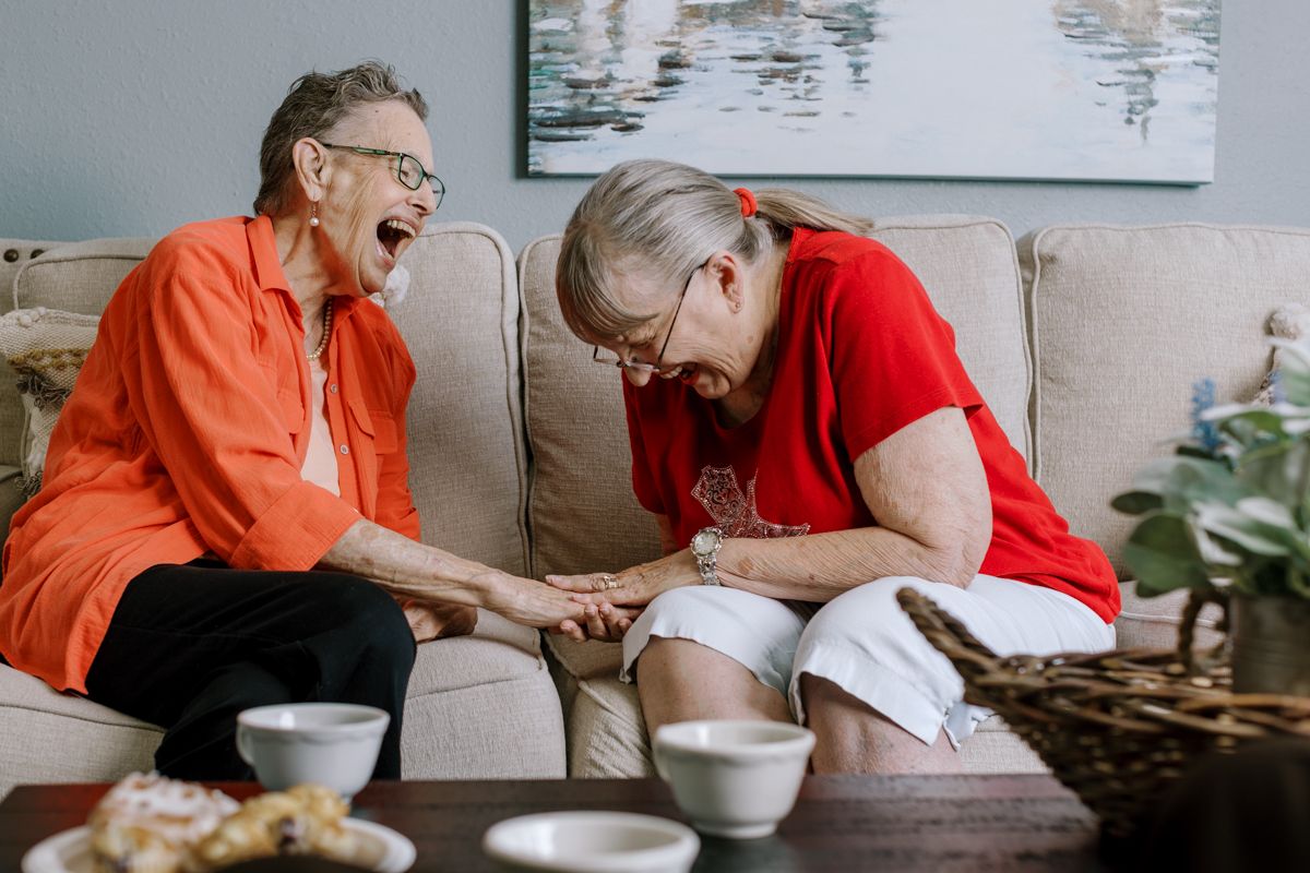 Two joyful elderly women laugh together while holding hands on a cozy couch in a senior living home.