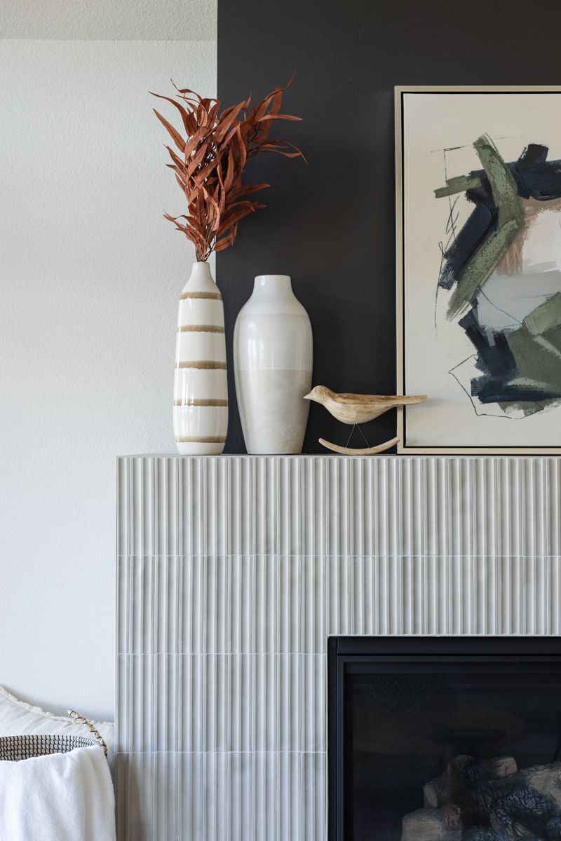 Modern fireplace with vertical tile surround, white ceramic vases, dried botanicals, and abstract art on dark wall.