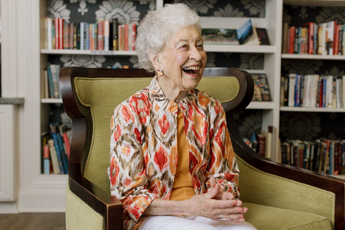 Happy elderly woman laughing in green armchair with colorful ikat jacket in front of bookshelf.
