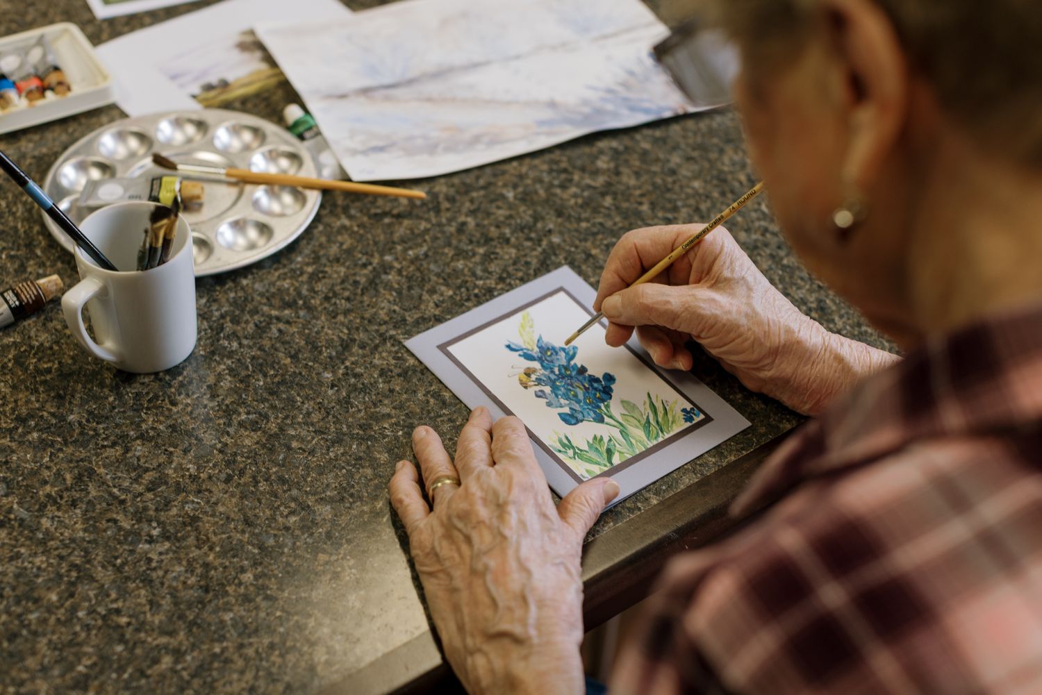 Elderly woman painting a detailed watercolor miniature artwork at a desk with brushes and a paint palette nearby.