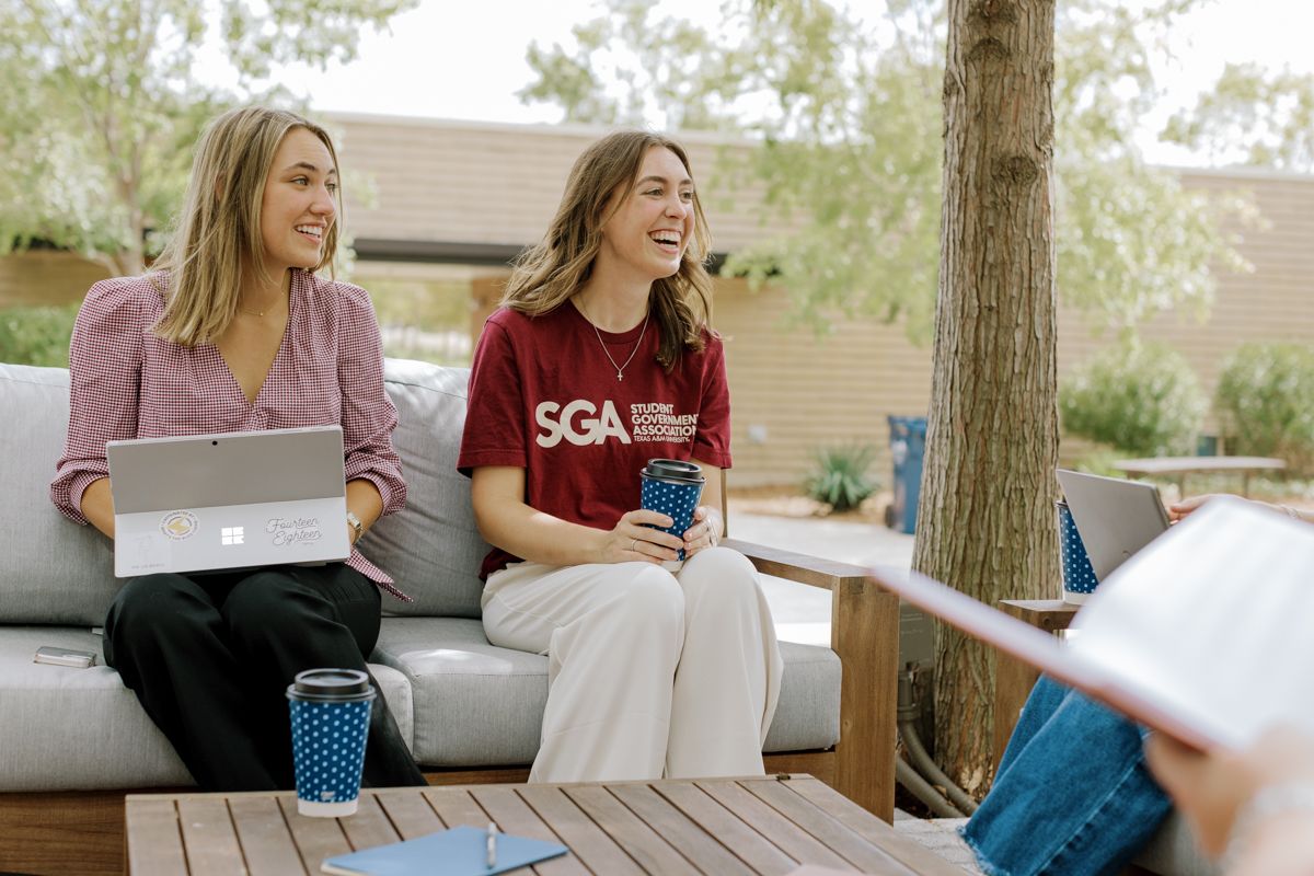 Two smiling college women relaxing on outdoor patio furniture, one with a laptop and one wearing an SGA t-shirt.