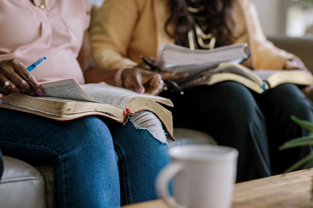 Two women sitting on a couch studying open Bibles together, taking notes, with a coffee cup on the table.