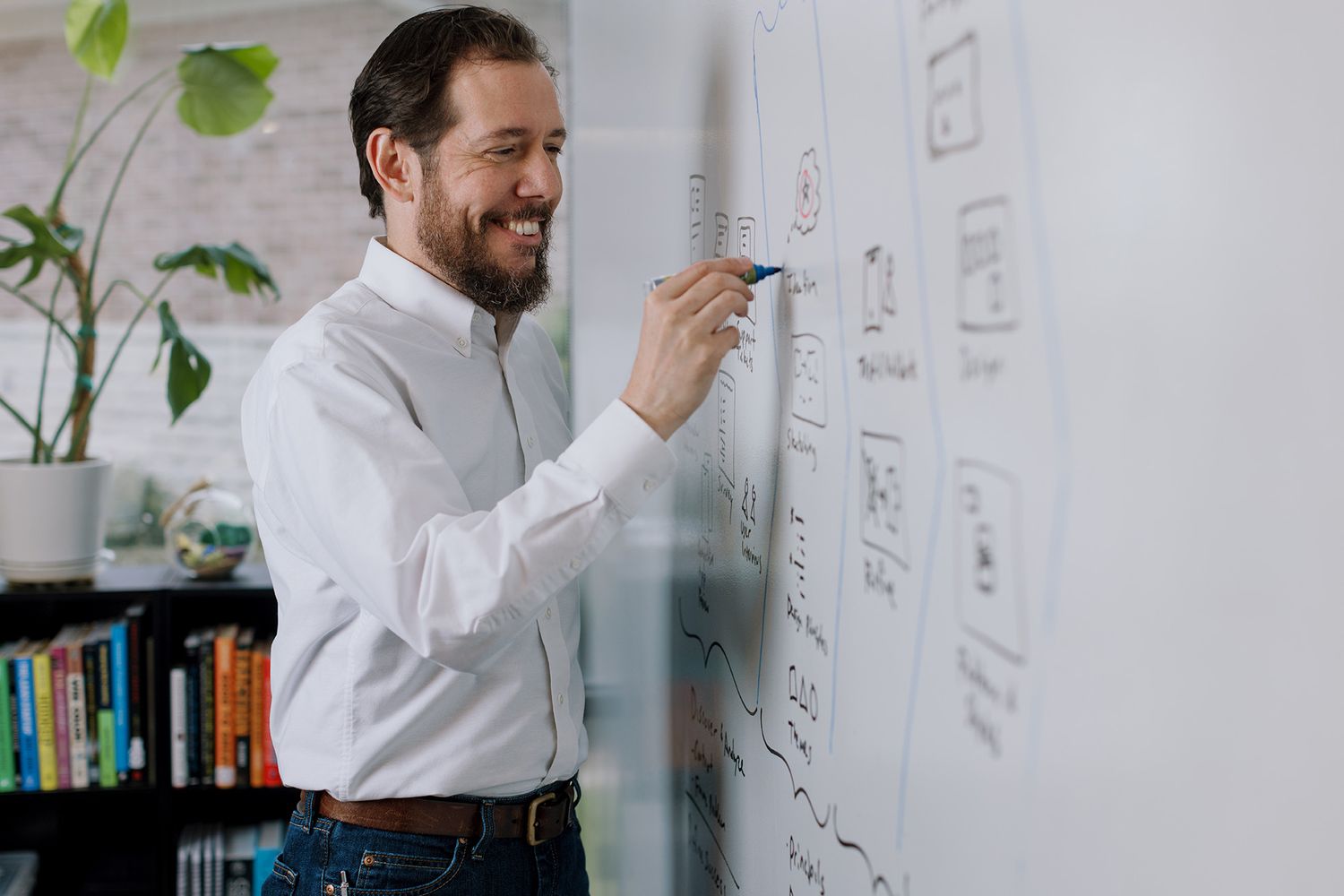 Smiling bearded man in white shirt writing on a whiteboard in a modern office with plants.