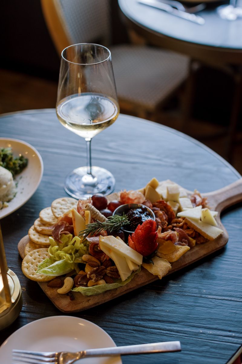 Charcuterie board with cheese, meats, crackers, and olives served with a glass of white wine on a dark table.