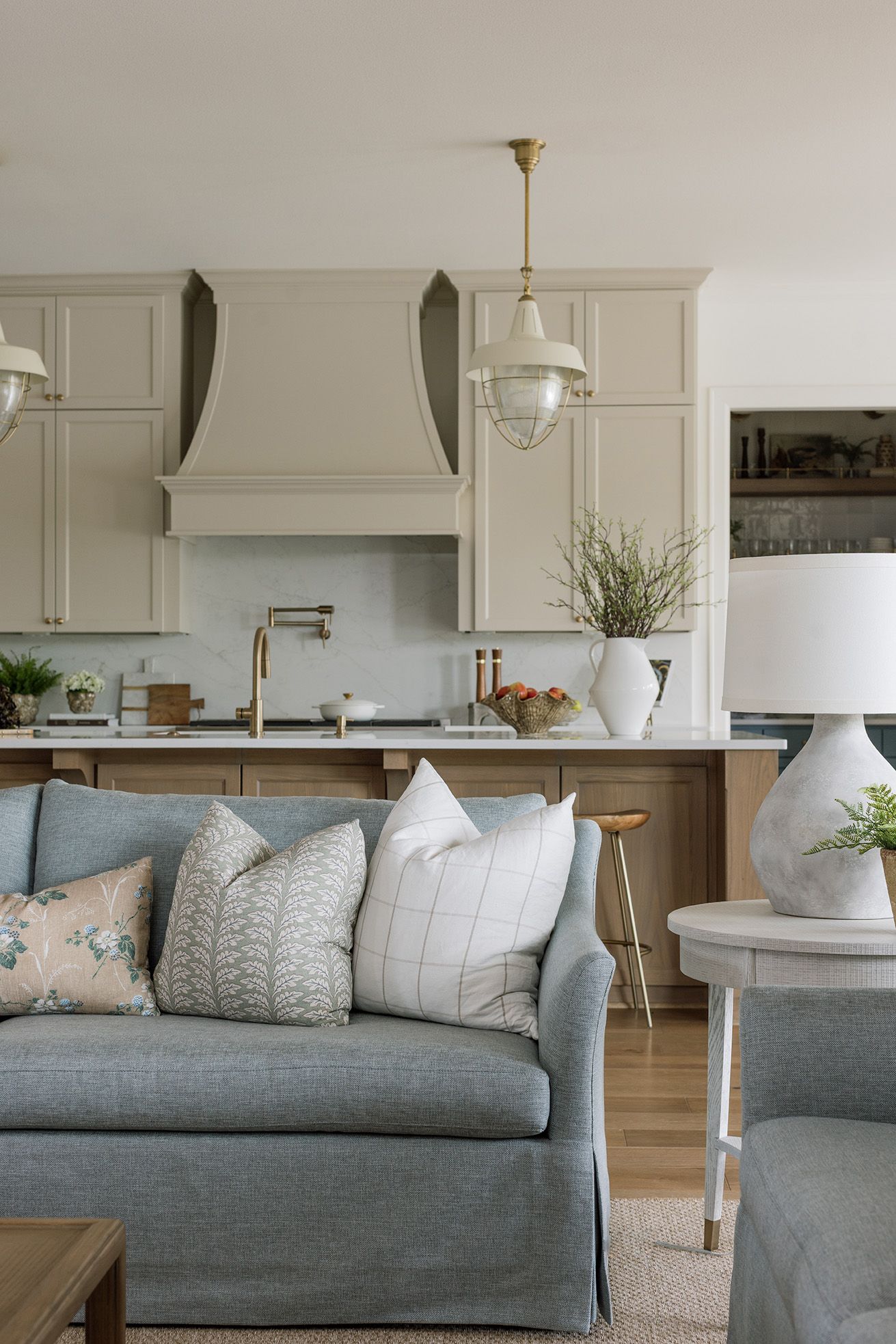 Open-concept living room with blue linen sofa and mixed pillows overlooking a taupe kitchen with brass pendant lights.