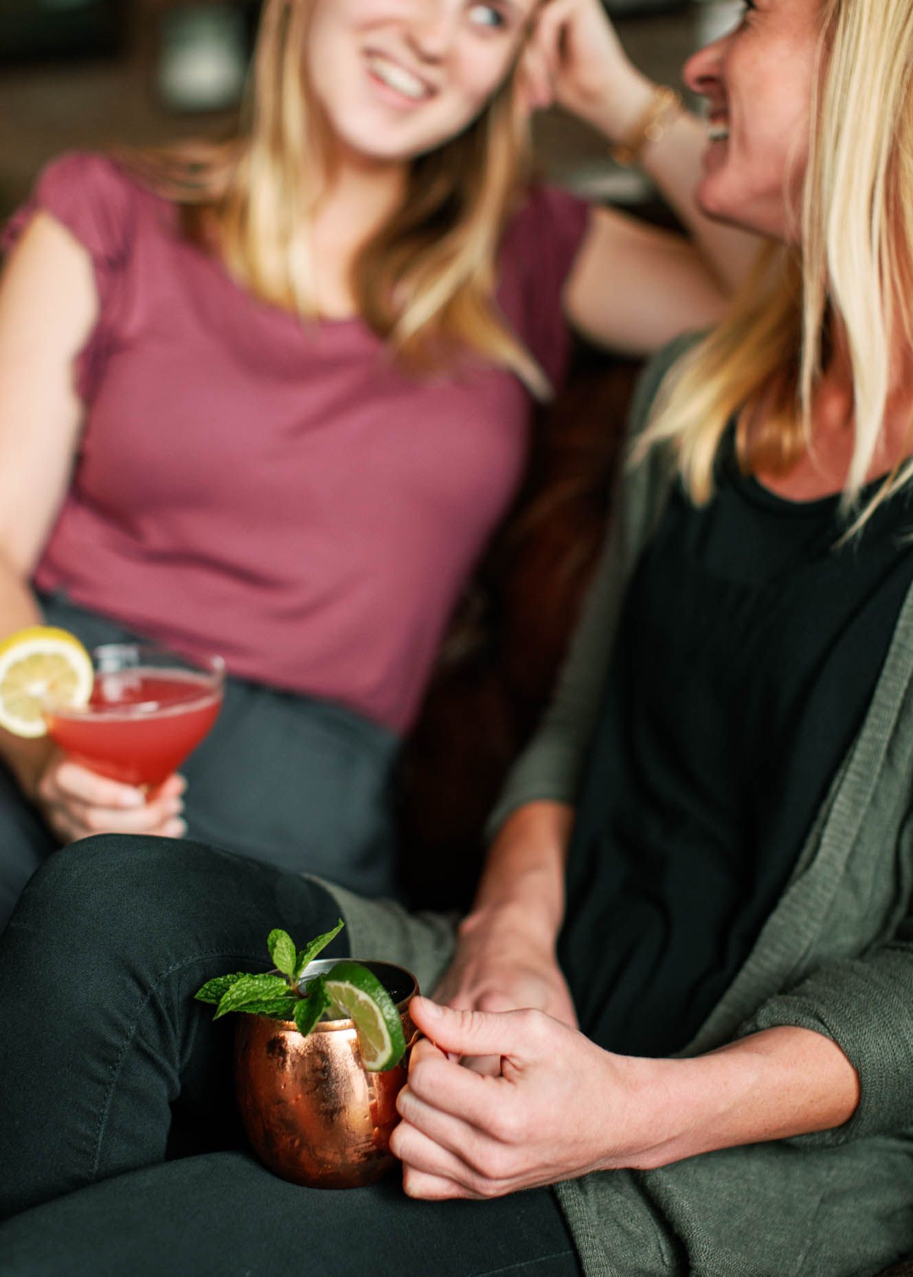 Two women socializing on a couch, holding cocktails including a Moscow Mule in a copper mug and a pink martini.