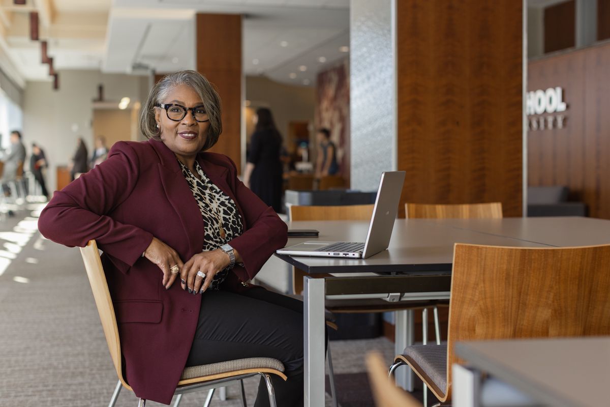 Confident older woman in burgundy blazer sits at a table in a modern school lobby.