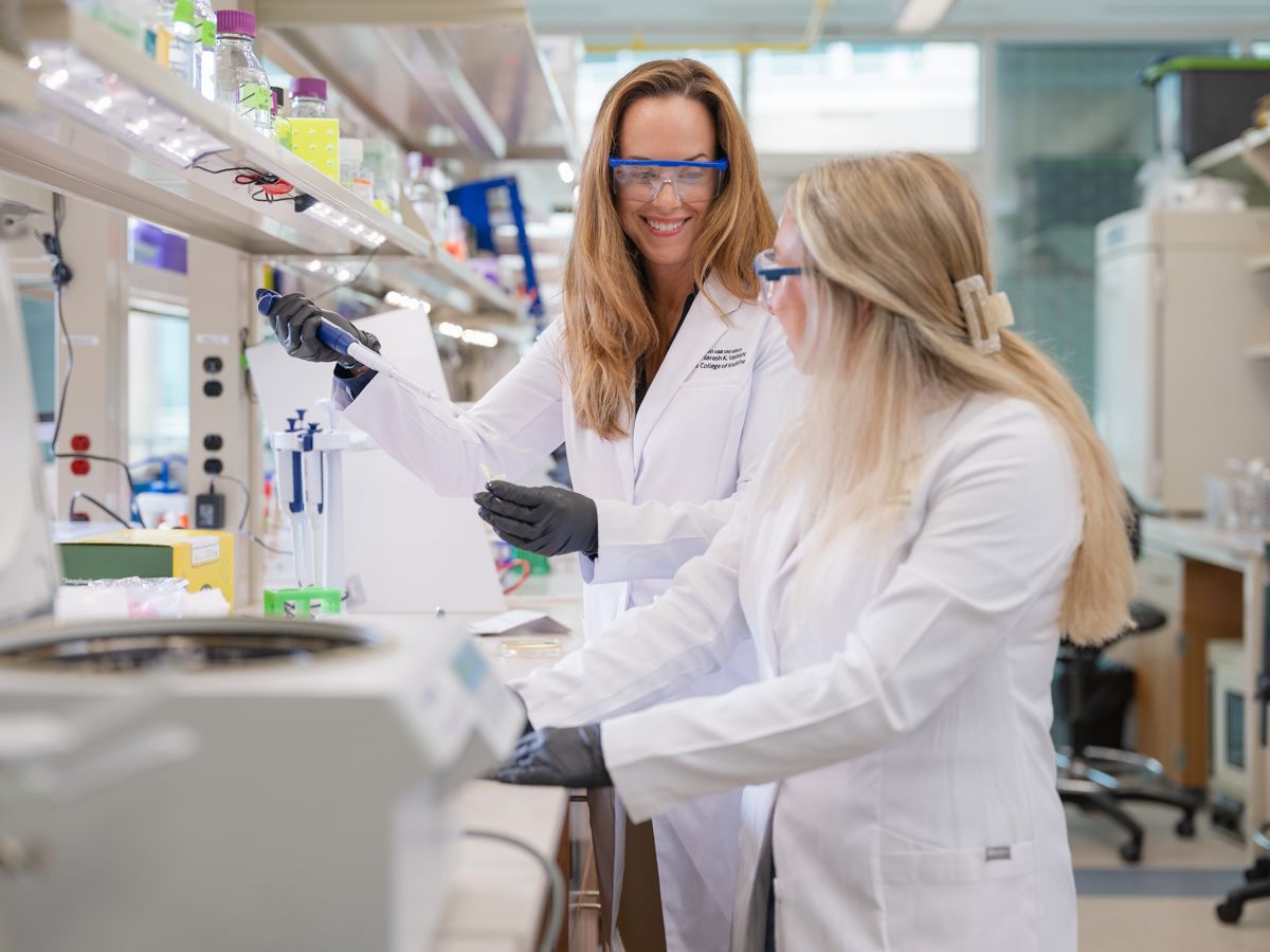Two female scientists in white lab coats and safety goggles working together in research laboratory.