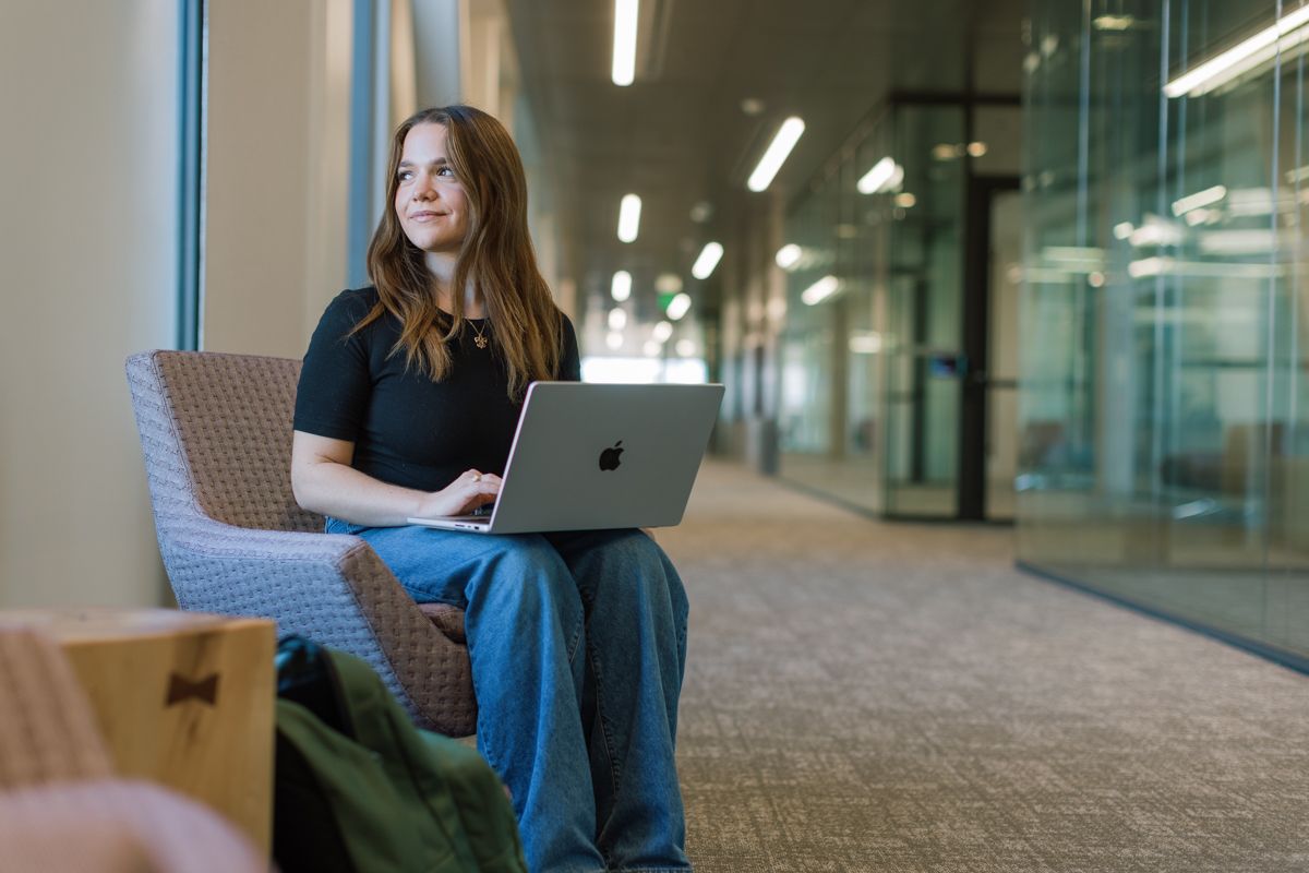 Female student sitting in a hallway using a MacBook laptop in a modern university building.