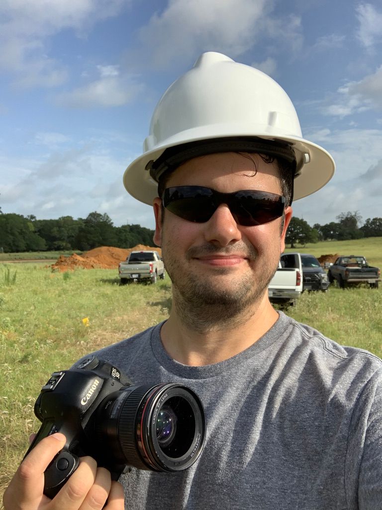 Man in white hard hat and sunglasses holds a Canon camera on a sunny construction site.