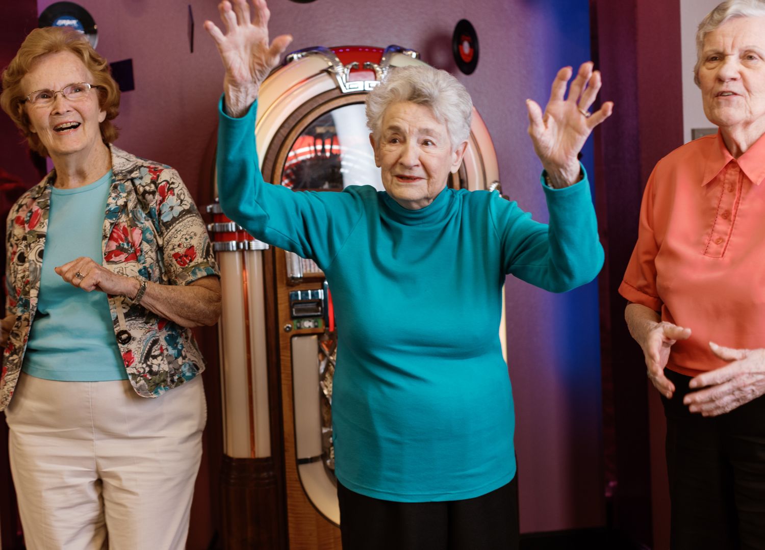 Three elderly women dancing joyfully near a vintage jukebox, raising their hands in celebration.