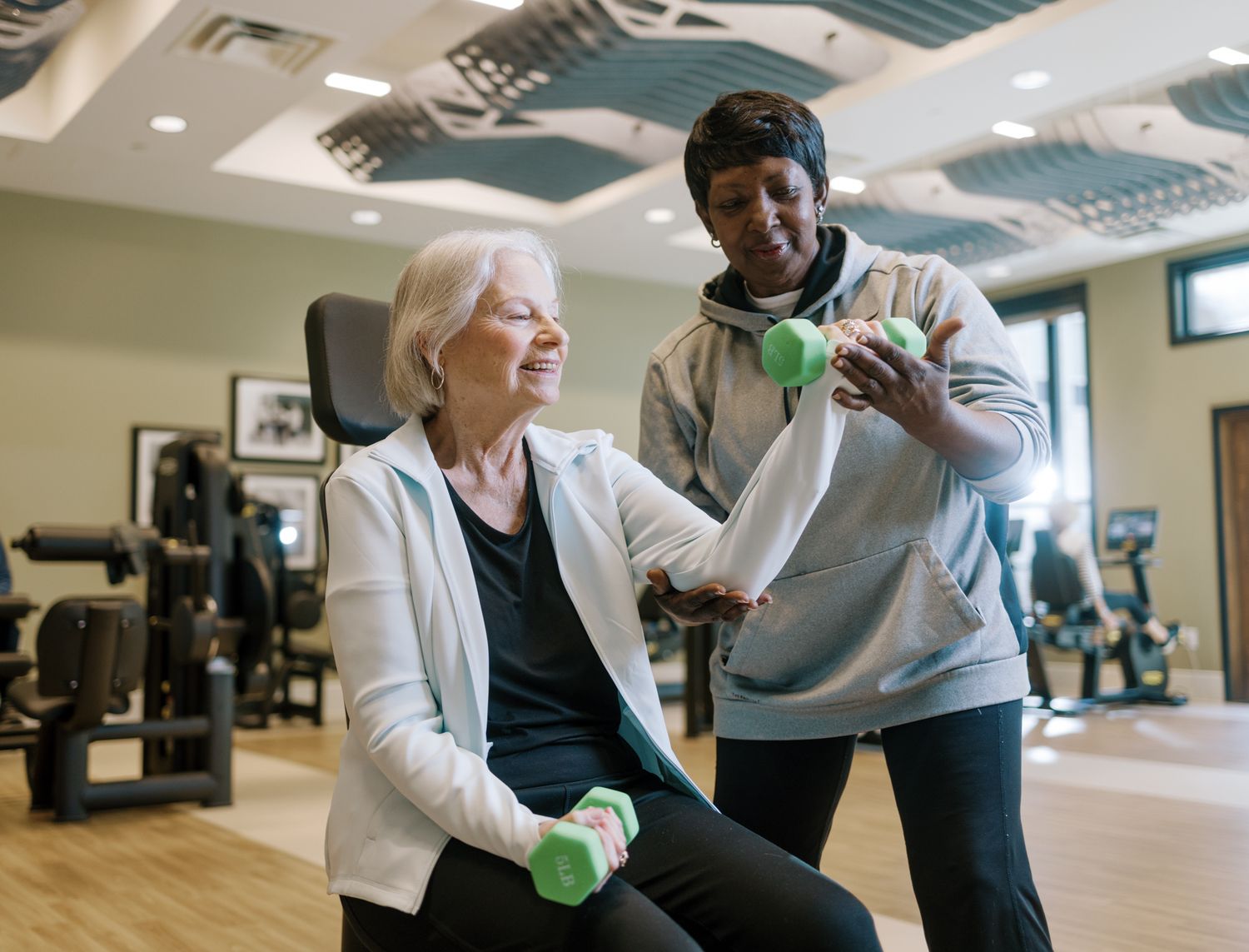 A personal trainer assists an elderly woman with dumbbell exercises in a bright, modern gym facility.
