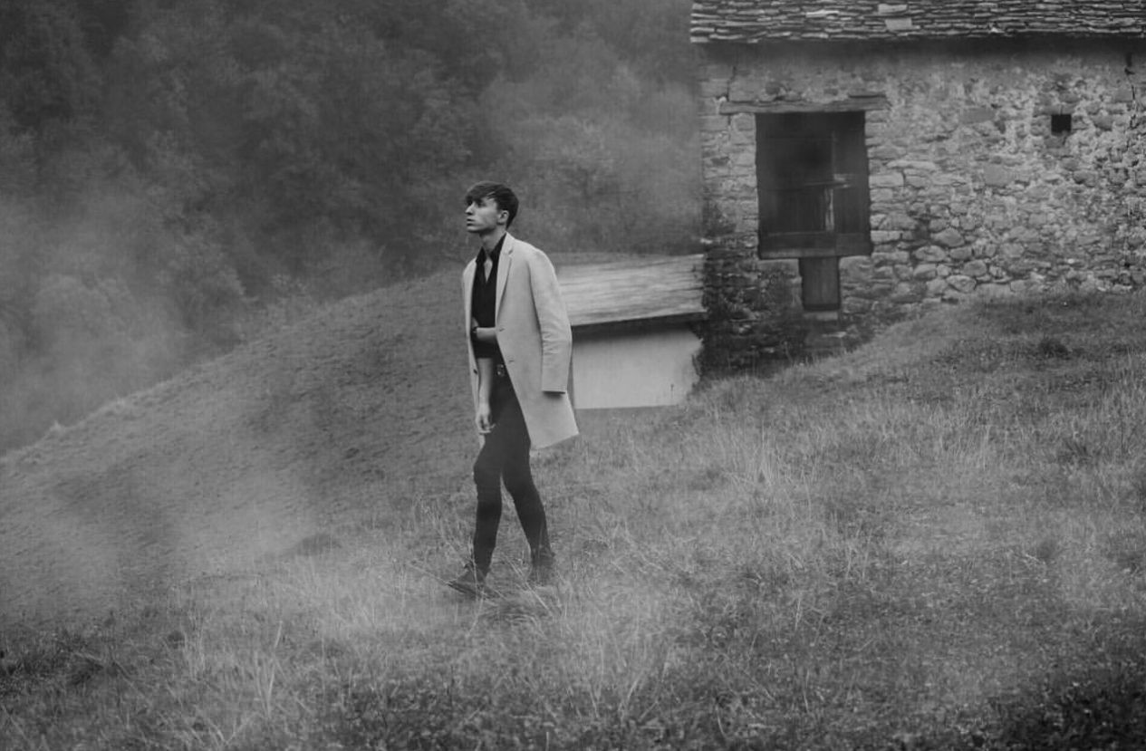 Man in long coat walking through misty field beside rustic stone building in black and white.