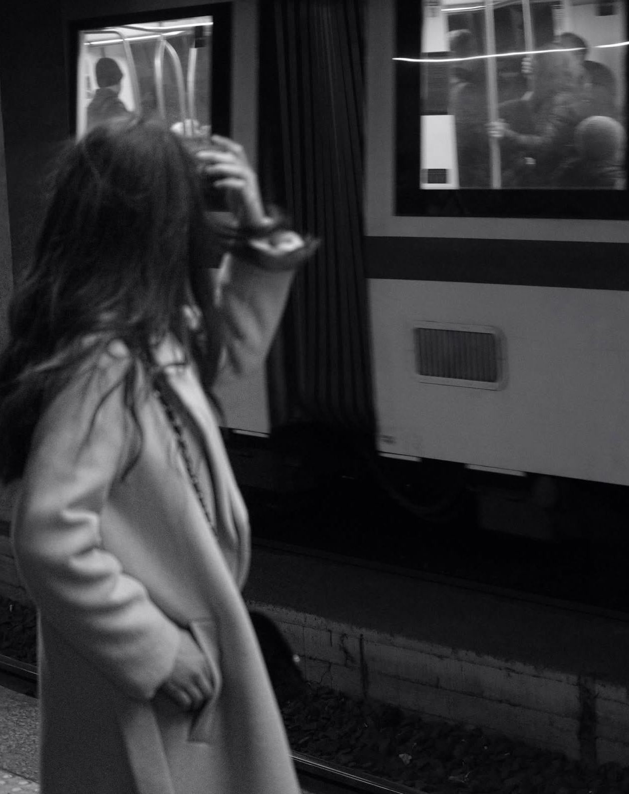 Black and white photo of a woman in a coat standing on a subway platform beside a train.