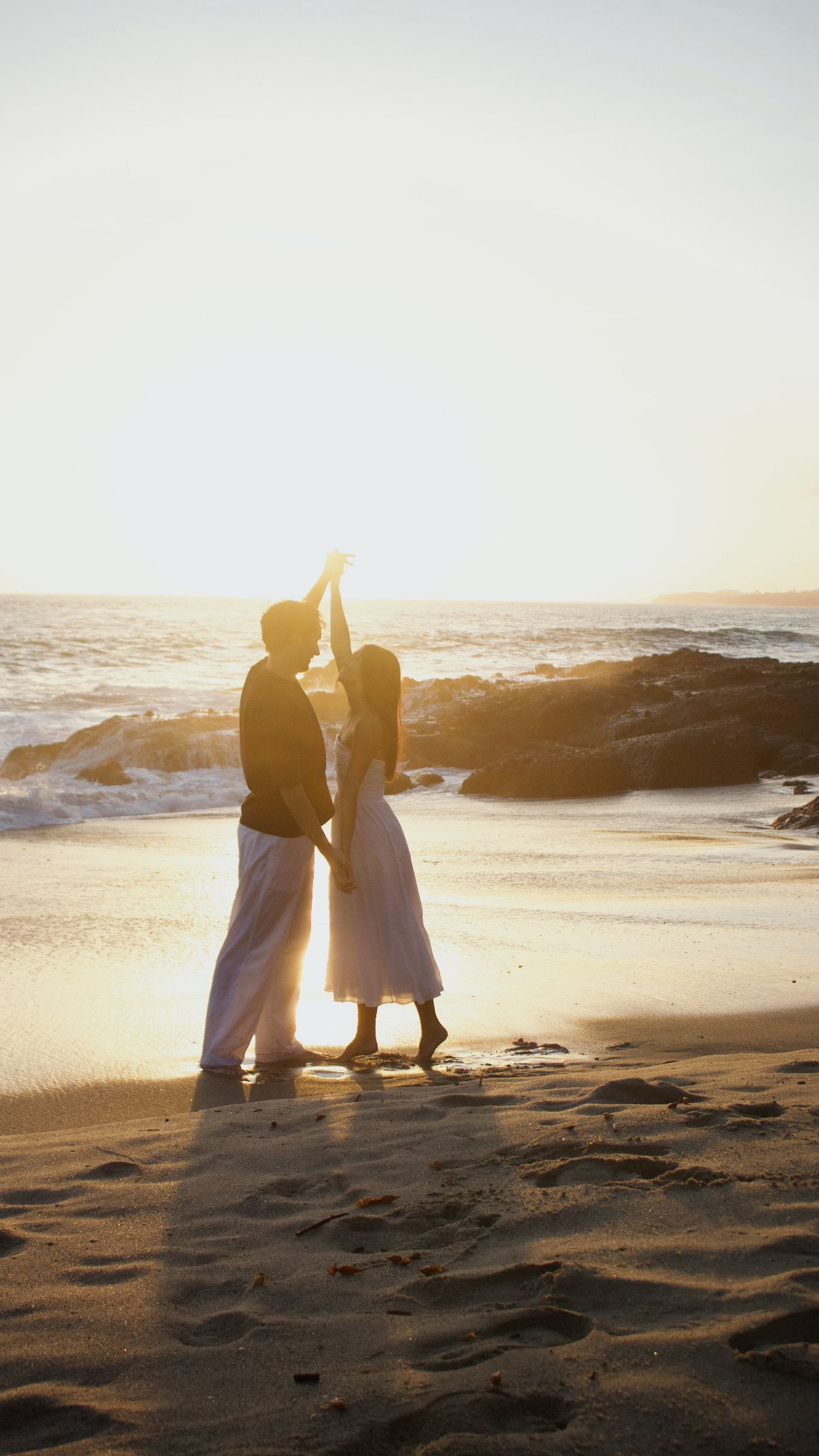 Couple dancing silhouetted on sandy beach at sunset with ocean waves and golden light.