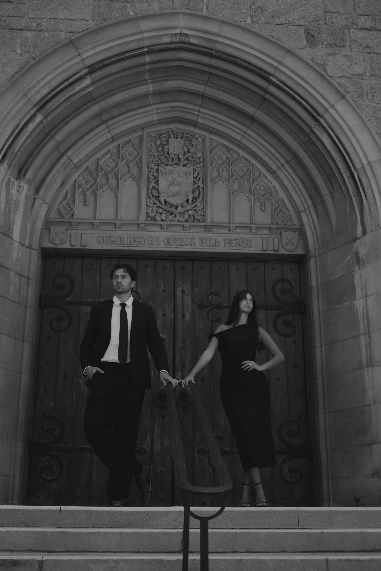 Black and white photo of an elegant couple holding hands before a grand arched stone church doorway.