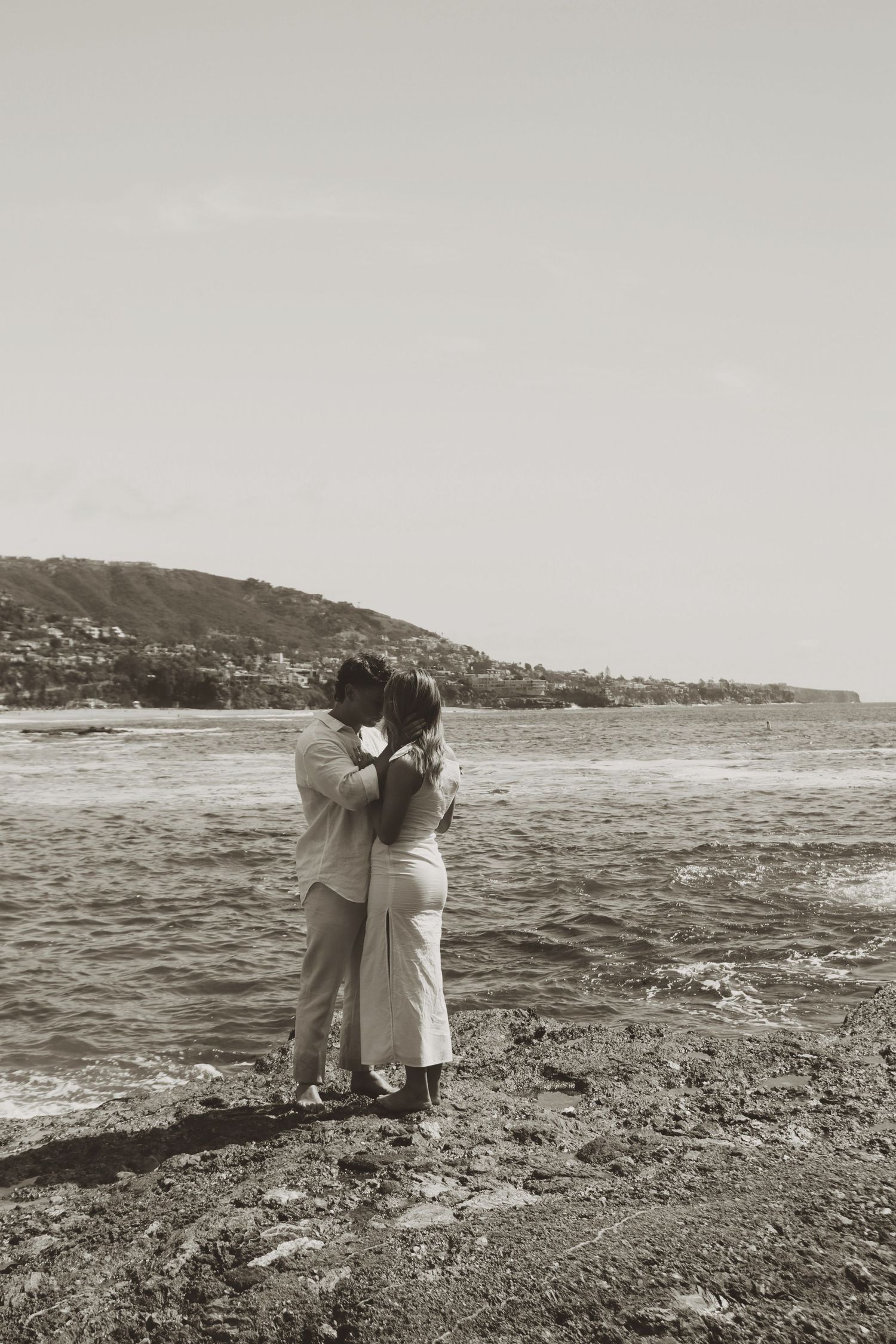 A couple shares a romantic kiss by the ocean shore in a black and white beach photo.