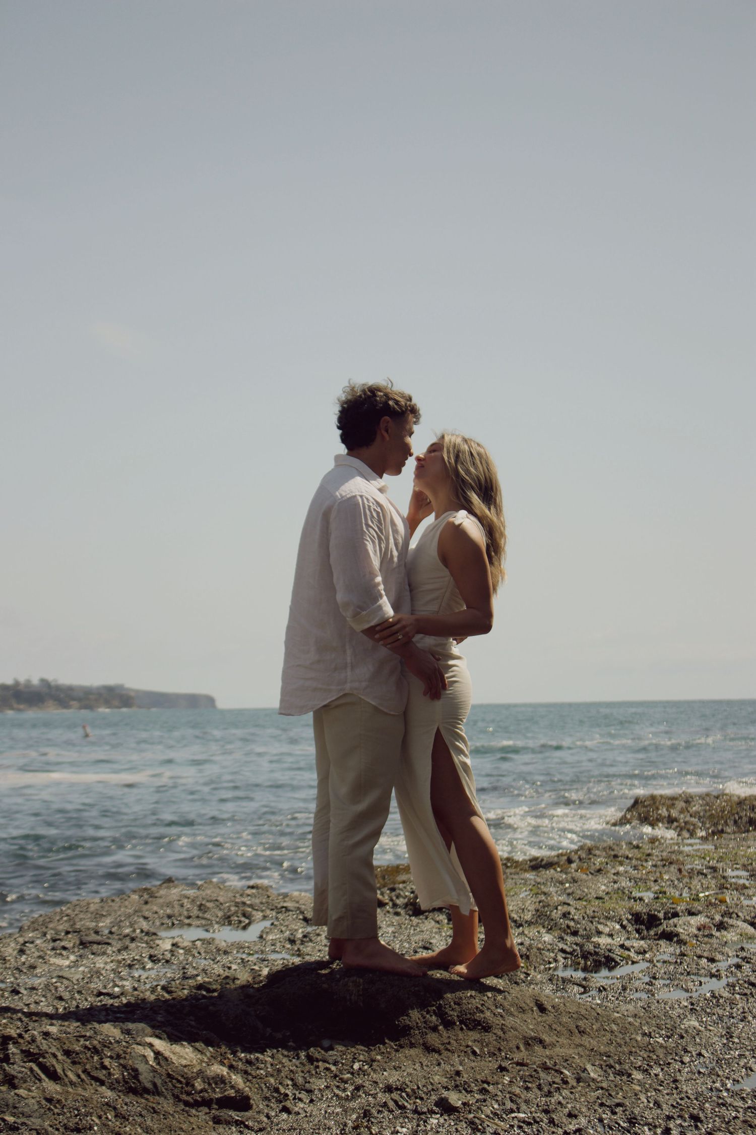 Romantic couple standing close together on rocky shoreline with ocean and clear sky behind them.