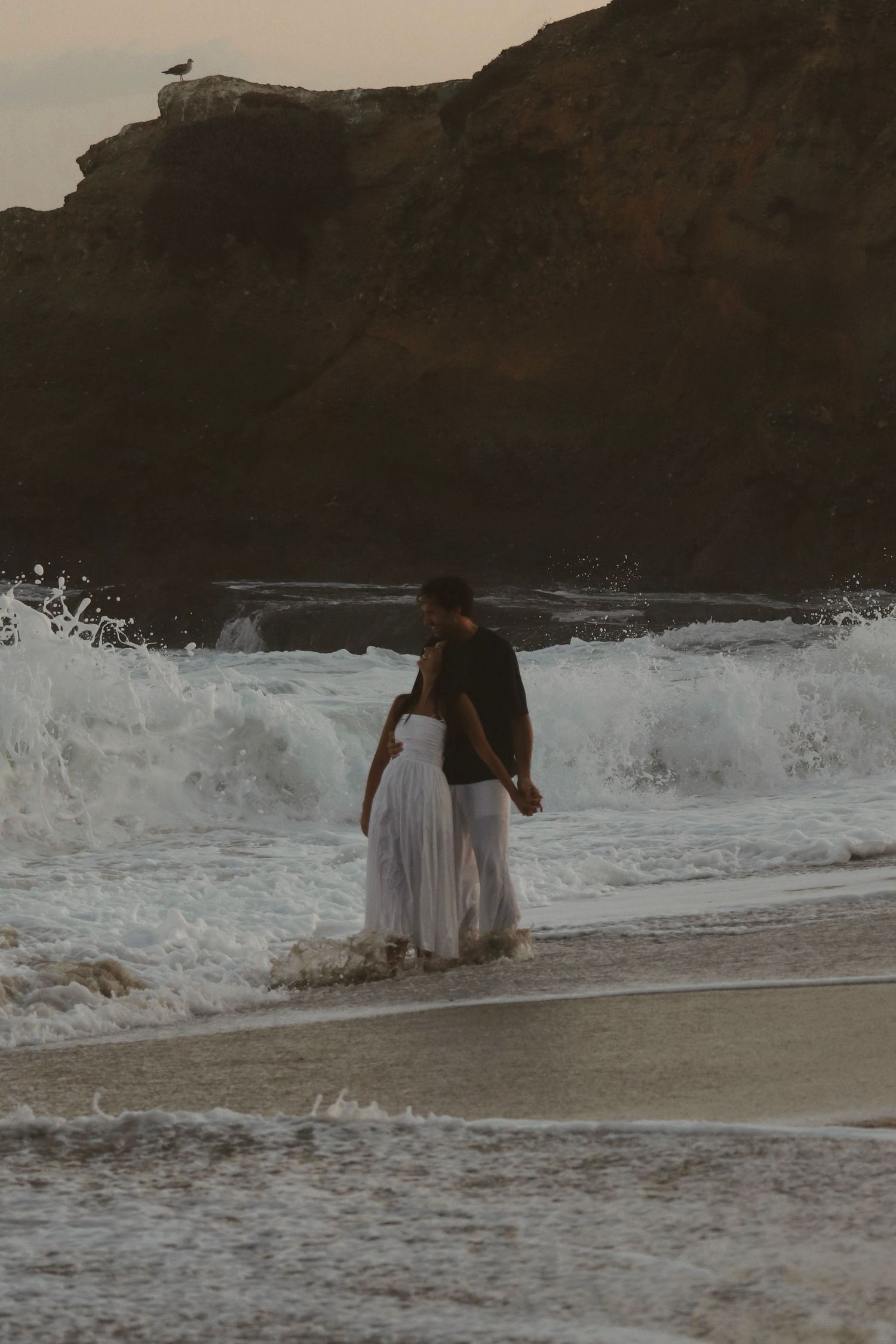 Couple in white dress and black shirt embracing at rocky beach shore with crashing waves at dusk.