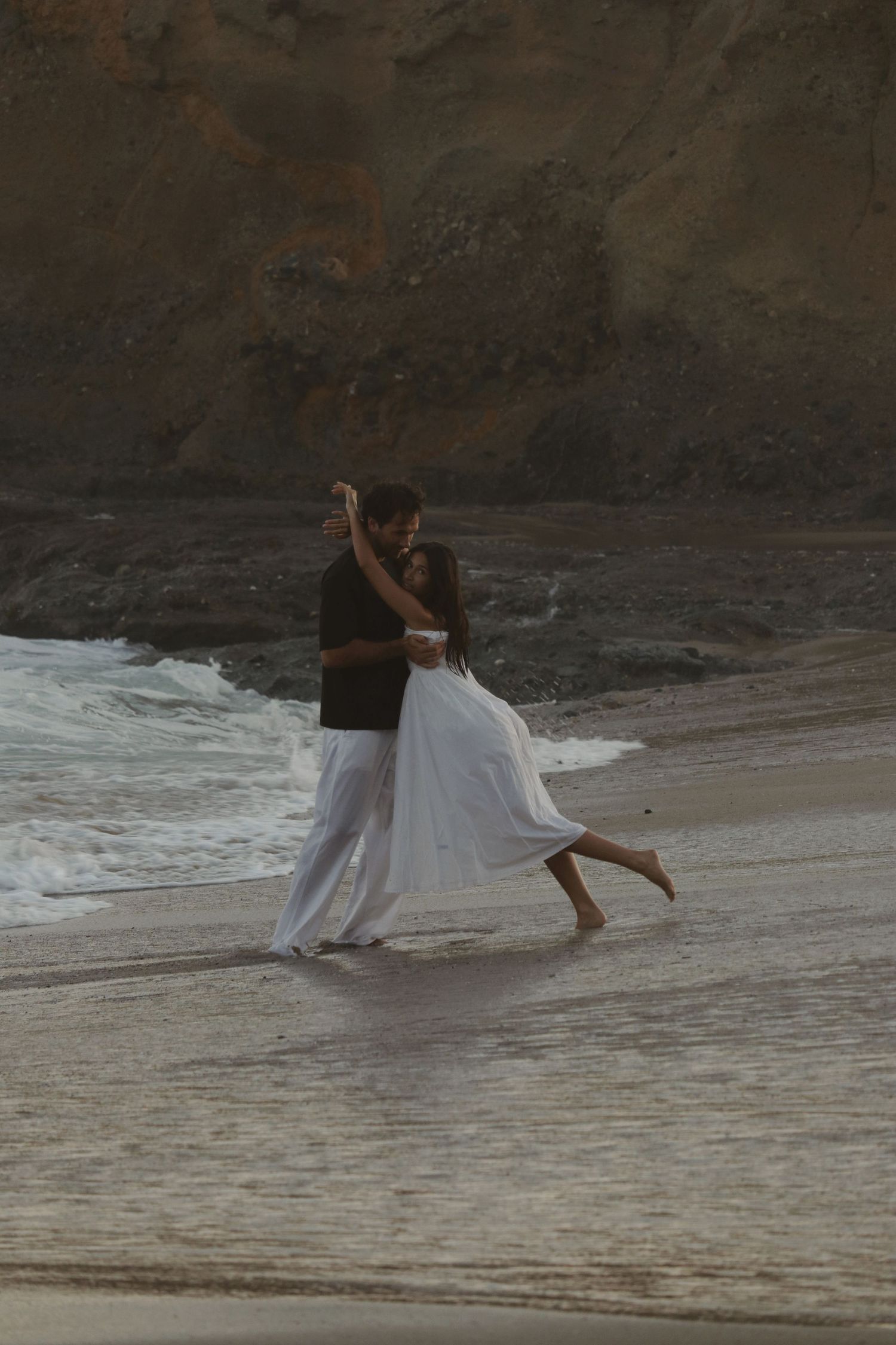 Romantic couple dancing barefoot on a sandy beach shore with ocean waves and rocky cliffs in the background.