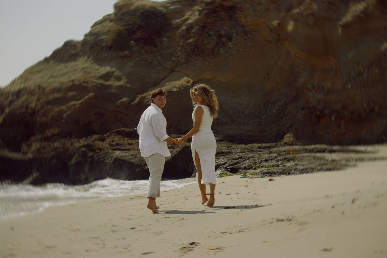 Couple in white outfits holding hands and dancing on sandy beach near rocky coastal cliffs.