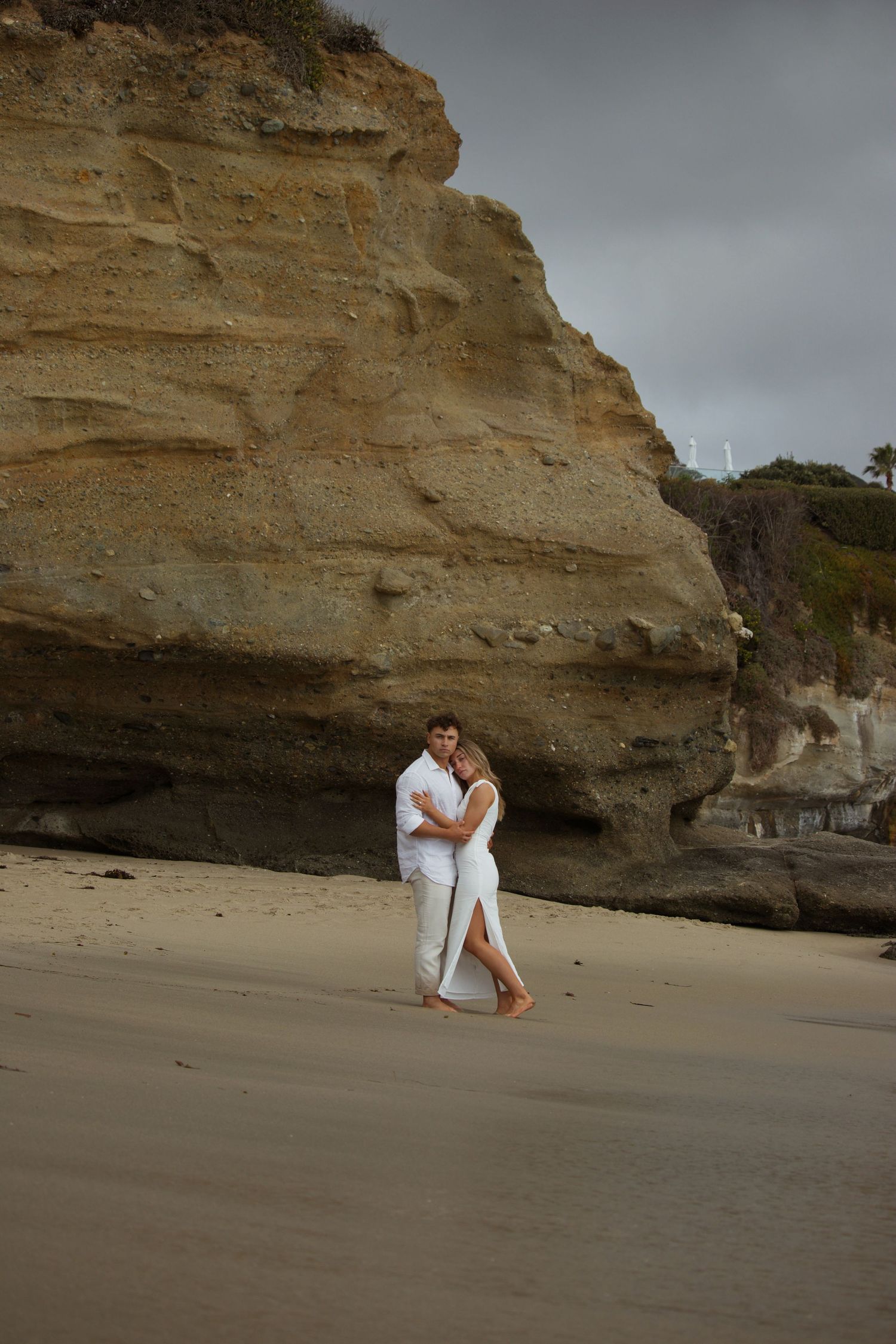 Couple embracing on sandy beach with dramatic sandstone cliffs backdrop, both dressed in white.