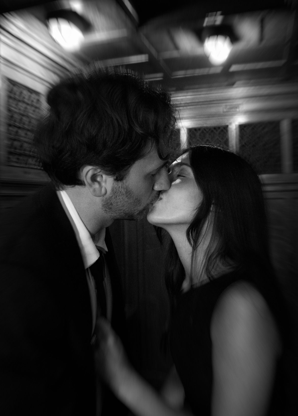 Black and white photo of a romantic couple sharing a kiss in a dimly lit elegant venue.