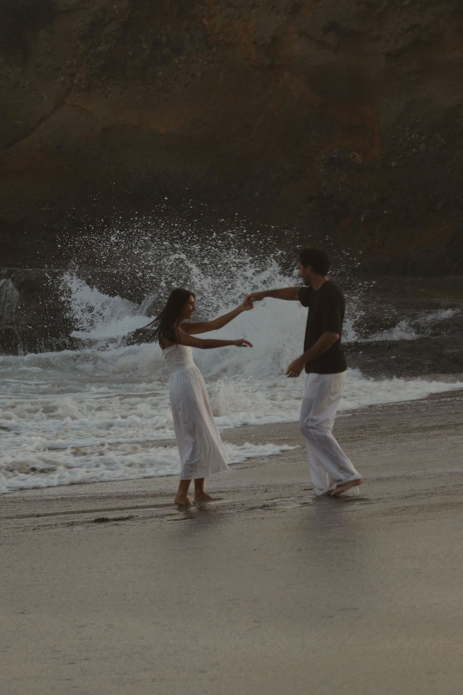Couple dancing romantically on a beach with crashing waves and rocky cliffs in the background.