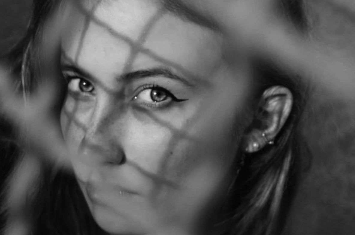 Black and white portrait of a young woman with long hair, viewed through a chain-link fence pattern.