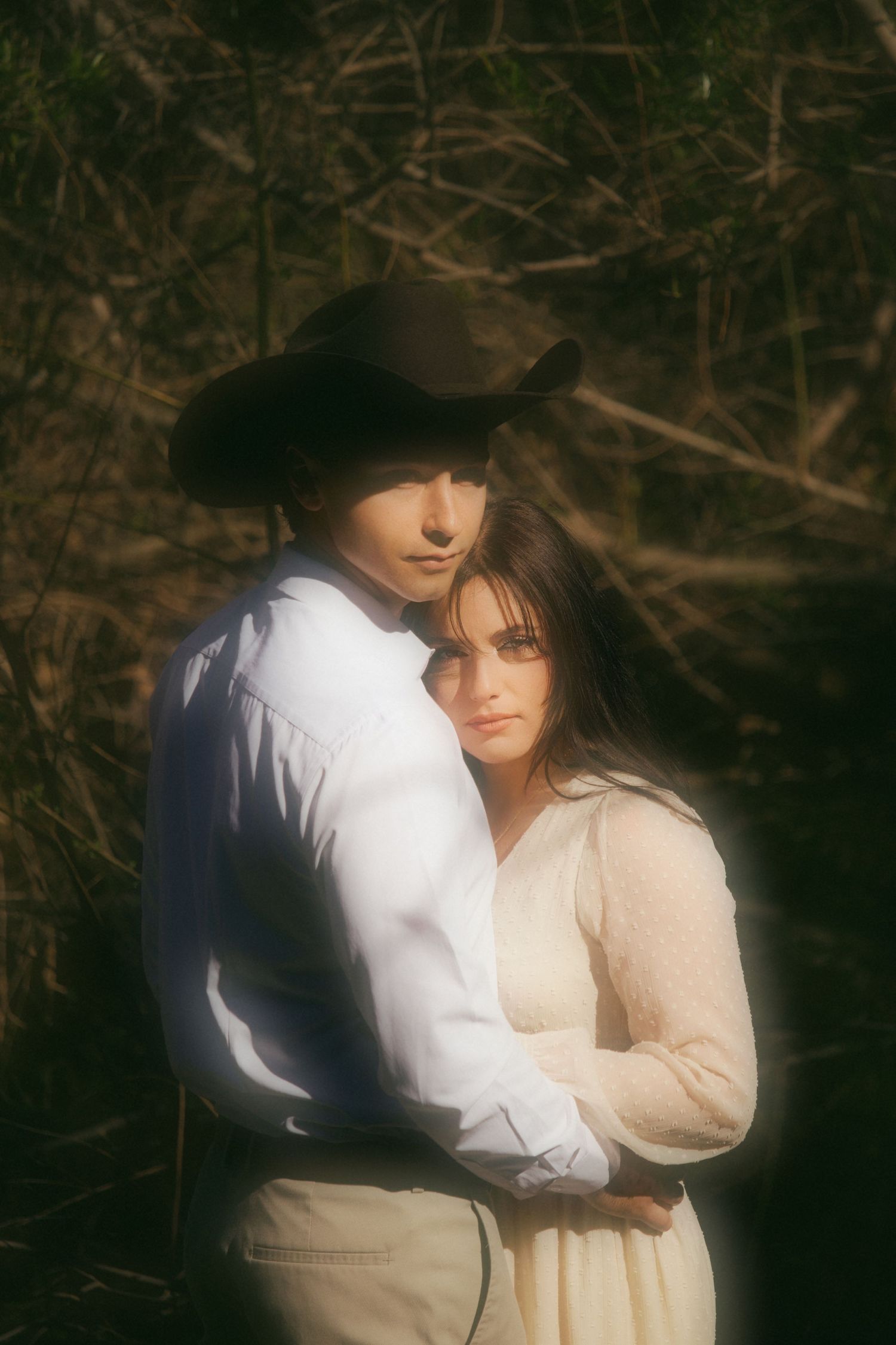 Couple embracing in warm golden light, man wearing cowboy hat and white shirt with woman in cream dress.